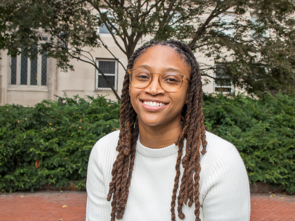 Black woman wearing glasses smiling at camera with trees and building in background